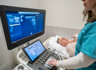 A patient lies on a bed receiving an ultrasound, while a technician operates the machine and monitors results on a large screen.