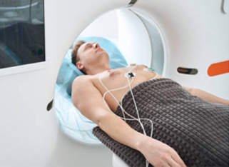 A patient lies on an exam table covered with a blanket, undergoing a CT scan, with electrodes attached to his chest