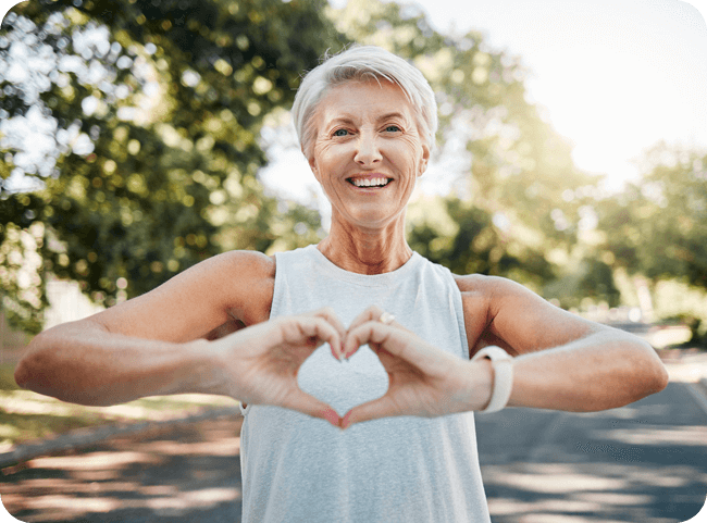 An elderly woman in a sleeveless shirt forms a heart shape with her hands, standing outdoors surrounded by greenery.