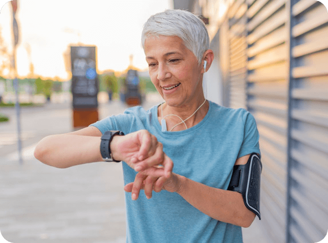 An older lady in a blue t-shirt checks her smartwatch while wearing earbuds, set against a city street at dawn.
